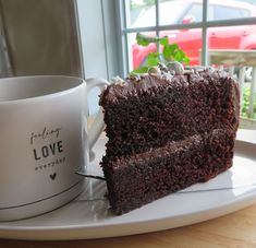 a slice of chocolate cake on a plate next to a coffee cup and window sill