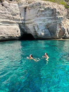 two people swimming in the blue water next to a large rock formation with a cave behind them