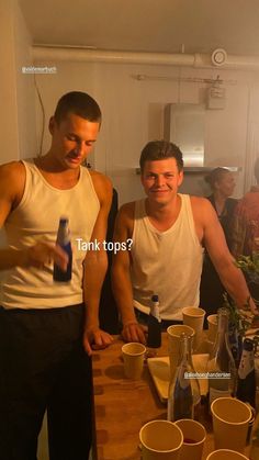 two men standing next to each other at a table with bottles and cups on it