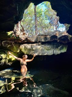 a woman is standing in the water near some rocks and trees with her arms outstretched