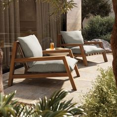 two wooden lounge chairs sitting next to each other on a stone patio surrounded by greenery