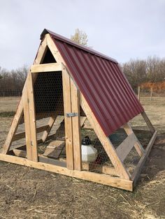 a chicken coop with a red roof and two chickens in the back ground, on top of dry grass