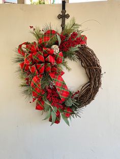 a christmas wreath hanging on the wall with red and green bows, greenery and a cross