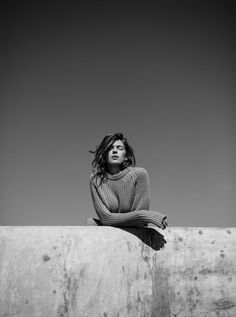 a woman sitting on top of a cement wall with her arms crossed and looking off to the side