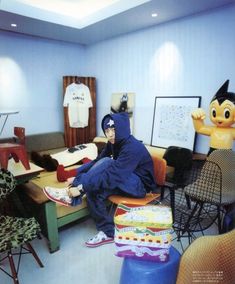 a man in a blue hoodie sitting on a couch surrounded by furniture and stuffed animals