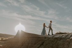 a man and woman holding hands walking on top of a hill with the sun in the background