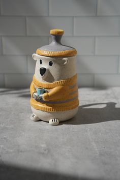 a ceramic bear with a sweater on holding a cup in it's paws and sitting next to a brick wall