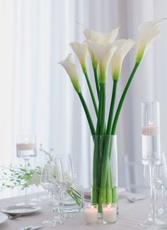 white flowers in a glass vase on a table with candles and wine glasses around it