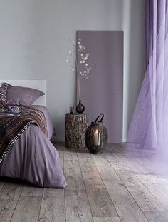 a bed with purple comforter and pillows in a room next to a vase filled with flowers