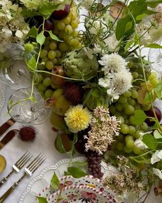 an arrangement of flowers, grapes and other greenery sits on a table set for dinner