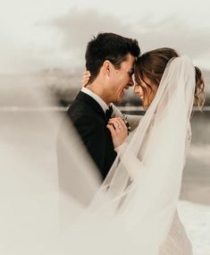 a bride and groom standing close to each other with veil blowing in the wind on their wedding day