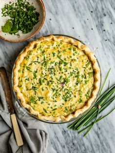 a quiche with cheese and green onions on a marble countertop next to a bowl of chives