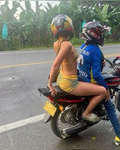 a man and woman riding on the back of a motorcycle down a street next to banana trees