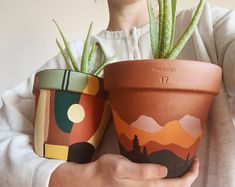 two potted plants with mountains and trees painted on them, one is holding the other