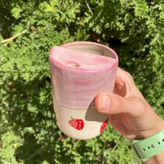 a person holding up a pink and white cup with strawberries on the outside in front of some bushes