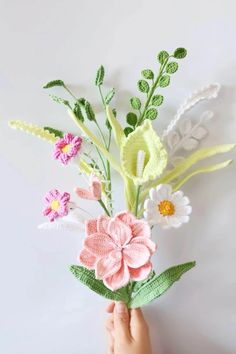 a hand holding a crocheted flower bouquet in front of a white wall with green leaves and flowers