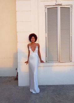 a woman standing in front of a window wearing a white dress and holding her hand on the wall