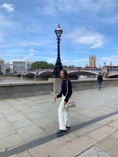 a woman standing on the sidewalk in front of a lamp post and bridge with people walking across it