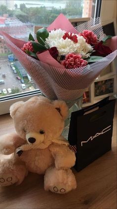 a brown teddy bear sitting next to a bouquet of white and red flowers in front of a window