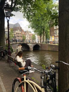 a woman sitting on a bench next to a river with bicycles leaning against the tree