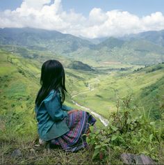 a woman sitting on top of a lush green hillside next to a valley with mountains in the background