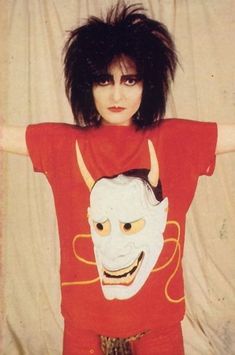 a woman wearing a red shirt with a demon mask on it's chest and her arms behind her head