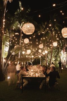 people are sitting at a table with lights hanging from the ceiling and around them is a palm tree