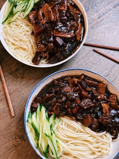 two bowls filled with noodles and meat on top of a wooden table next to chopsticks