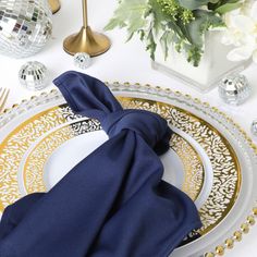 a white plate topped with a blue napkin and gold rimmed charger next to a vase filled with flowers