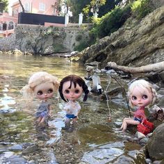 three dolls are playing in the water near some rocks and trees with houses in the background