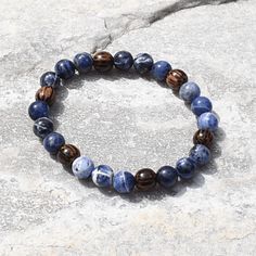 Experience the essence of harmony with the artisan-made Sodalite and Palm Wood Unisex Bracelet. This captivating accessory unites the tranquil allure of Sodalite and the rustic charm of Palm Wood, creating a wearable masterpiece that resonates with both elegance and earthy energy. Centered around the serene Sodalite beads, the bracelet showcases a palette of deep blues interwoven with delicate patterns, reminiscent of the vast expanse of a starlit sky. Sodalite is renowned for its ability to fos Earthy Blue Bracelets As Gift, Earthy Blue Bracelets For Gifts, Sodalite Beaded Bracelets With Natural Stones For Gift, Sodalite Bead Bracelets As Gift, Spiritual Sodalite Gemstone Beaded Bracelets, Sodalite Round Beads Bracelets As Gifts, Handmade Sodalite Round Bead Bracelets, Sodalite Round Beads Bracelets For Healing, Handmade Lapis Lazuli Bracelets For Meditation