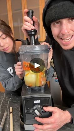 a man and woman standing in front of a blender filled with fruit, making a smoothie