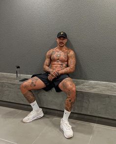 a man with tattoos sitting on a bench in front of a gray wall and wearing white tennis shoes