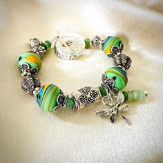 "This colorful chunky glass bead and gemstone bracelet is comprised of 14mm artisan Lampwork glass beads done in etched vibrant green, blue and golden yellow with black dots by glass artist Natalia Darlin. Each Lampwork bead is gently capped with Karen Hill Tribe Silver ornate bead caps, oxidized to show lovely pattern detail. A 1/2\" by 1/4\" flower patterned Karen Hill Tribe Silver cylinder focal bead sits in the center of the bracelet with a sterling Silver dragonfly charm and Chrysoprase charm dangling on the side, adding a unique look. ** It is said seeing dragonflies alive or as charms is symbolic of a loved one who has passed on from this earth** The bracelet is accented with 5mm Chrysoprase beads and Karen Hill Tribe Silver beads and spacer beads. I finished the bracelet with a lar Dragonfly Bracelet, Dragonfly Charm, Hill Tribe Silver, Boho Bracelet, Bracelet Boho, Lampwork Glass Beads, Vibrant Green, Black Dots, Bracelet For Women