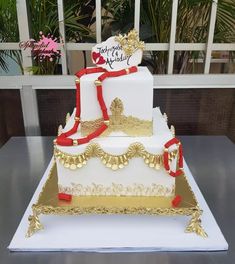 a white and gold wedding cake with red ribbon on it's side, sitting on a table