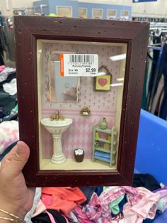 a person holding up a miniature dollhouse in a frame with clothes on the floor