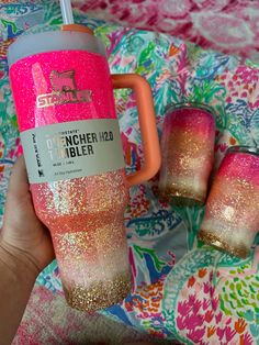 a person holding a pink and gold glitter tumbler next to two mugs on a colorful blanket