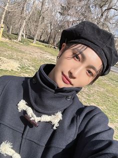 a young man wearing a black hat and coat in a park with trees behind him