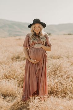 a pregnant woman wearing a brown dress and black hat standing in a field with her hands on her stomach