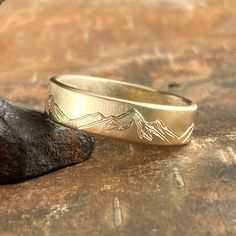 a gold wedding band with mountains engraved on the side, resting on a piece of wood