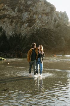 a man and woman are walking in the water near some rocks on a beach together