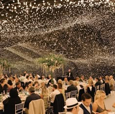 many people are sitting at tables with white tablecloths and lights hanging from the ceiling