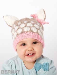 a baby girl wearing a knitted hat with a deer pattern on the side and a pink bow at the top