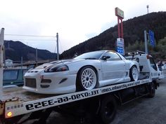 a white car is on the back of a flatbed tow truck with mountains in the background