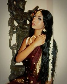 a woman with long black hair wearing a sari and gold jewelry sitting in front of a statue