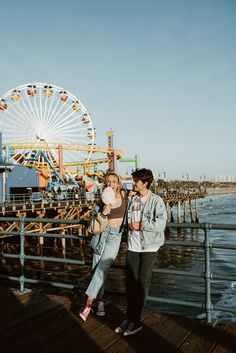 a man and woman standing next to each other on a pier near the ocean with a ferris wheel in the background