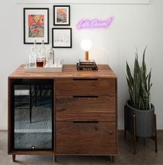 a wooden cabinet sitting next to a wall with pictures on it and a plant in the corner