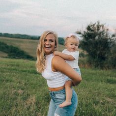 a woman holding a baby in her arms while standing in a field with trees and grass