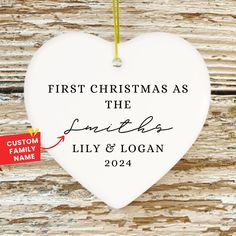 a white heart ornament hanging on a wooden wall with a red ribbon and the words, first christmas as the smiths lily & logan