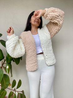 a woman standing next to a plant wearing white pants and a pink cardigan sweater